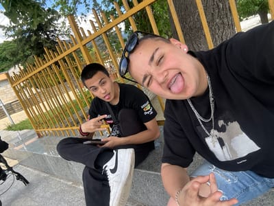 Two young people are posing for a selfie outdoors. The foreground subject, on the right, is wearing sunglasses on their head, a black T-shirt, blue ripped jeans, and multiple silver necklaces. This person has braces and is sticking their tongue out, winking, and making a hand gesture. The second person, in the background on the left, is sitting on a concrete ledge, wearing a black T-shirt, black pants, and white sneakers. They are also sticking their tongue out and making a hand gesture. Behind them is a yellow metal fence and a tree. A portion of a bicycle is visible in the lower left corner of the frame. The background reveals additional greenery and buildings, suggesting an urban or suburban park setting. The picture was taken during the day. It is likely that the location is Sopot, Bulgaria, because the question mentions this location. However, without additional information or context from the image, specific features confirming that the location is in Sopot, Bulgaria cannot be determined.