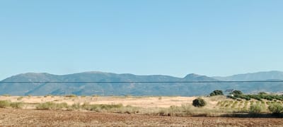 This image depicts a rural landscape with a clear blue sky. In the foreground, a tilled field of reddish-brown soil is visible. Beyond this field, a dry, grassy plain stretches out, dotted with sparse green shrubs and trees. A thin black horizontal line, likely a wire, traverses the middle of the image. In the mid-ground, a cluster of trees and rows of young plants are present, with a small building and solar panels situated on a slight elevation. The background is dominated by a range of low, rolling mountains covered in muted green vegetation under the vast expanse of the sky. The scene is likely located in the municipality of Lucillos, Spain, given its characteristic dry, agricultural landscape and mountainous horizon. No people or animals are visible in the frame.
