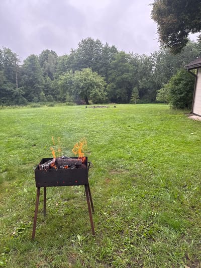 The image depicts an outdoor scene in a grassy area, likely a backyard or park, with a small charcoal grill in the foreground. The grill contains burning wood and logs, with flames reaching upwards. The surrounding area is covered in green grass and scattered with some weeds. In the background, there is a dense line of trees forming a forest. To the right of the image, a portion of a building with a white exterior and a brown roof is visible. The sky is overcast, suggesting a cloudy day. The specific location in Antakščiai, Lithuania is not discernible from the visual elements alone. There are no people or animals present in the frame.