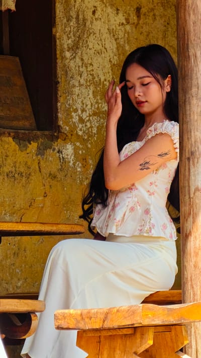 A young Asian woman with long dark hair is sitting on a wooden bench in front of a textured, weathered yellow wall. She wears a white floral top and white skirt. Her right hand is raised near her forehead. A tattoo of three birds in flight is visible on her upper left arm. The background suggests a rustic setting, possibly in Lũng Cú Commune, Vietnam, given the architectural style and environment. The lighting indicates an outdoor daytime scene. The woman is situated on a wooden structure that appears to be part of a building or sheltered area.