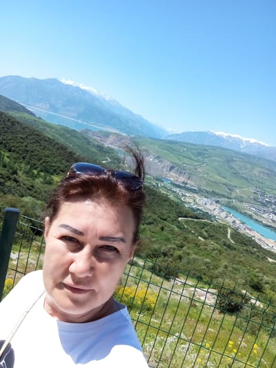 The image shows a woman taking a selfie in Chinarkent, Uzbekistan. She is standing on a hillside overlooking a valley with a turquoise river running through it. Snow-capped mountains are visible in the background. The woman has dark hair, sunglasses on her head, and is wearing a white t-shirt. A metal fence is in the foreground. The weather appears sunny.