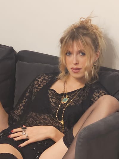 A fair-skinned woman with blonde hair pulled up in a messy bun sits on a grey couch, facing the camera. She wears a sheer black lace top, a gold necklace with a turquoise pendant, and black thigh-high stockings. Several rings adorn her fingers, which rest on her lap. The background is a plain, light-colored wall. There are no indications of Pantin, France in the image.
