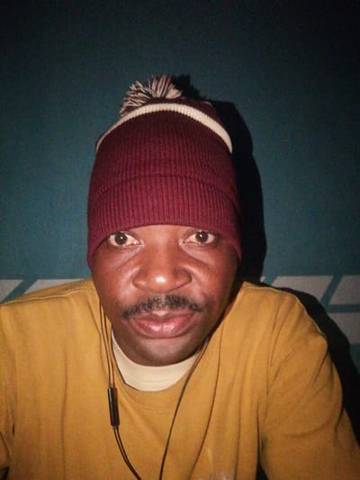A close-up, head-on selfie of a Black man wearing a maroon beanie with a white and maroon pom-pom and a yellow t-shirt with a light-colored undershirt. He has a mustache and appears to be wearing earbuds. The background is a dark teal wall with some lighter geometric patterns visible at the bottom. The lighting is dim, with a shadow cast on the right side of his face and shoulder. The image appears to have been taken indoors in a dimly lit room, likely in Ajasse Ipo, Nigeria, given the context.