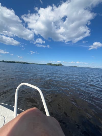 The image depicts a view from a boat on a body of water, likely a large lake or river, with a distant tree-lined shore under a blue sky filled with white, puffy clouds. In the foreground, a white railing and a portion of a person's arm and knee are visible, suggesting the perspective of someone on the boat. The water is dark and rippled, indicating gentle movement. The shoreline is densely populated with trees. The scene is set during the daytime, with bright sunlight illuminating the sky and water. Without specific landmarks or identifying features of Cheboksary, Russia, the exact location within the city cannot be determined from this image alone.