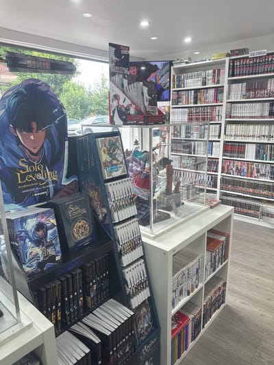 The image shows the interior of a manga and comic book store, likely located in Villeneuve-d'Ascq, France, as indicated by the presence of the store name "LE PETIT JAPON" visible on a sign above the entrance. The store is filled with bookshelves organized by genre, featuring a wide selection of Japanese manga.
In the foreground, a large promotional display for "Solo Leveling" is prominent, featuring artwork of the main character. Below this, several volumes of "Solo Leveling" manga are neatly stacked and displayed. To the right, a glass display case contains anime figurines, including what appears to be a character holding a hammer and shield, and another with elaborate colorful hair.
The shelves in the background are densely packed with manga volumes. A section labeled "SEINEN" (a category of manga targeted at adult men) is clearly visible on one shelf. Other shelves showcase various manga series, with spines showing titles and volume numbers. A television screen in the upper left displays an anime or manga-related program. The store has a clean, modern aesthetic with white shelving and a light-colored wooden floor. The exterior of the store is visible through large windows, showing trees and a street with a parked car, suggesting a typical urban or suburban setting.