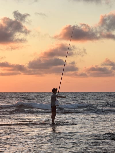A lone person stands in the shallow water at the edge of the ocean, holding a long fishing rod. The person is silhouetted against a warm, orange and pink sky filled with scattered clouds, indicative of a sunset. The ocean is slightly choppy with small waves rolling towards the shore. A white bucket hangs from the fishing rod. The scene appears to be the coast of Haifa, Israel, based on the provided context.