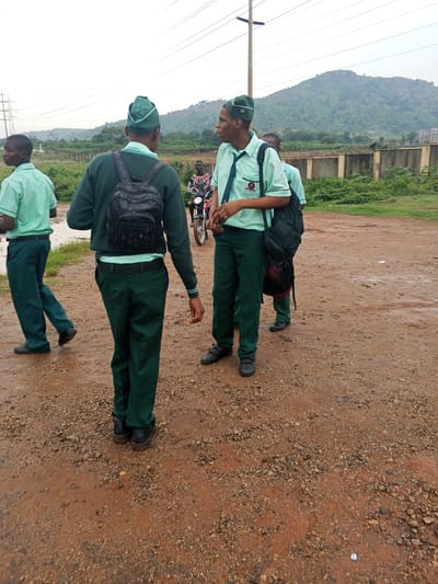 Three young males, likely students, are standing in a dirt area that appears to be a road or pathway. They are all wearing matching green uniforms with collared shirts and dark green trousers. Two of them have backpacks, one black and one partially visible. One of the students is wearing a green cap.
In the background, a motorcycle is visible with a person riding it. Beyond that, there is a wall or fence and lush green vegetation. In the far distance, a large, rocky, and verdant hill dominates the landscape under an overcast sky. Power lines are also visible stretching across the sky. The ground is damp, suggesting it may have recently rained or is a wet area.
The students appear to be in conversation or waiting for something. Their expressions are neutral. The time of day appears to be daytime, given the ambient light. The location is suggested to be Nigeria due to the landscape features and the presence of a motorcycle. There is no visible text in the image.