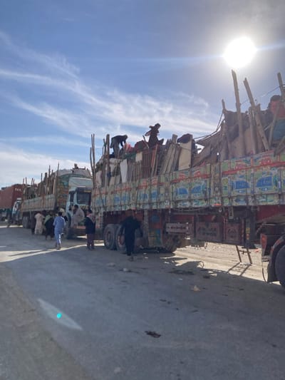 The image depicts a daytime scene in Chaman City Tehsil, Pakistan, featuring a queue of multiple heavily loaded cargo trucks on an unpaved or gravel road under a bright blue sky with wispy white clouds and a prominent sun.
The foreground is dominated by the right rear of a large, multi-axle truck adorned with elaborate, colorful "truck art" decorations. This truck's bed is piled high with a diverse assortment of cargo, including what appears to be wooden planks, bundled textiles, and various household items or furniture, secured with ropes and tarpaulins. Two individuals are positioned on top of this overloaded cargo, actively manipulating or adjusting items. A third individual is partially visible atop cargo on a truck further back.
To the left, a line of several similar, decorated trucks extends into the distance. Near the cab of the second truck in the line, approximately five to seven men, dressed in traditional attire (shalwar kameez or similar tunics), are gathered on the ground. Another man in a light blue tunic walks away from the camera between the first and second trucks. In the immediate foreground, a man dressed in dark clothing walks towards the front of the nearest truck.
The road surface appears rough and dusty, with shadows indicating strong sunlight from the upper right of the frame. The scene suggests a busy transit or loading area.