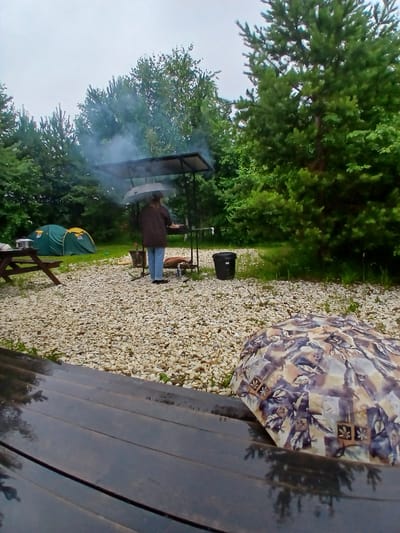 The image depicts a campsite in Verkhneye Dubrovo, Russia. A person is standing under an umbrella near a barbecue pit, with smoke rising from it. Two green tents are set up in the background. A wooden picnic table is visible on the left side of the image. The ground is covered with gravel and some patches of grass. A patterned umbrella lies folded in the foreground. The environment is surrounded by trees, suggesting a natural setting. The sky is overcast, indicating potentially rainy weather.