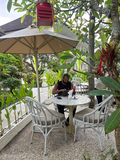 In Karangasem, Indonesia, a man with a beard sits at a round marble-topped table outdoors, in what appears to be a cafe or restaurant setting. He is wearing a black sleeveless shirt and is focused on a dark green pouch he is holding. Two white wicker chairs with beige cushions surround the table. A red candle in a glass holder sits on the table, along with a white napkin and a phone. In the background, lush green foliage and flowering plants create a tropical ambiance, with a large umbrella providing shade over a portion of the seating area. The ground is covered in small, light-colored pebbles. The architecture in the distance suggests buildings common to the region.