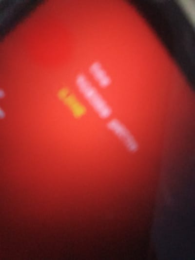 The image depicts a close-up, blurry view of a red surface with some indistinct white and yellow shapes. The red surface appears to be a reflective material, possibly a screen or a glossy object, with light glinting off it. The lighting is uneven, with brighter areas and darker shadows. The overall impression is abstract due to the blurriness and lack of clear focus on any specific subject. There is no discernible information about the location in Ilorin, Nigeria, or any specific details about people, objects, actions, or interactions.