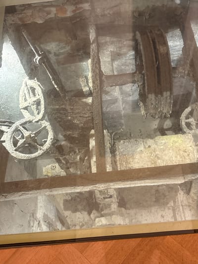 This image shows a section of an old mechanism, likely part of an industrial or water-powered system, housed within a stone structure. Prominently featured are two large, circular wooden wheels with spokes, heavily coated in dust and grime, suggesting disuse. One wheel is positioned above and slightly behind the other, and they appear to be part of a pulley or winch system, possibly with rope remnants. There are also substantial wooden beams and structural elements supporting the machinery, all showing signs of age and deterioration. The environment appears to be an interior space, possibly a basement or exhibition area, with visible stone walls and floor. The scene is viewed through a glass pane, as indicated by reflections and glare on the surface. This appears to be a historical exhibit, possibly located in Gatchina, Russia, given the context.