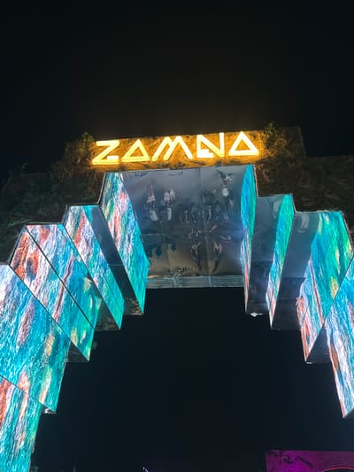 The image depicts a unique architectural display at night. The structure is a series of ascending platforms or steps, seemingly made of reflective material displaying a vibrant, swirling blue and orange pattern suggestive of water. In some areas, the reflective surfaces below the patterned platforms show a crowd of people. The structure is attached to a textured vertical column that appears to be illuminated by an adjacent, vertically-oriented neon sign spelling "ZAMANA" in yellow. The environment surrounding the structure is dark, suggesting a nighttime setting. The presence of the "ZAMANA" sign and architectural style suggests the location is Giza, Egypt.