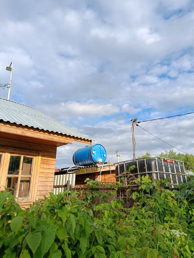 This media file depicts an outdoor scene in a rural setting, likely a backyard or garden area in Verea, Russia. The main subjects are a wooden structure with a corrugated metal roof, resembling a small house or shed, and two large water tanks. One is a bright blue barrel, and the other is a black IBC tote with a metal cage. Lush green foliage, possibly raspberry bushes and other wild plants, dominates the foreground, suggesting a summer or late spring setting.
The time of day appears to be late afternoon or early evening, indicated by the soft, diffused sunlight and the way it illuminates the clouds and the side of the wooden building. The weather is partly cloudy, with a mix of blue sky and fluffy white clouds.
There are no people visible in the image. The activity taking place is not explicit, but the presence of water tanks suggests their purpose is for water storage, possibly for gardening or household use. Notable details include a streetlamp or utility light pole near the wooden building and electrical wires running across the sky, supported by a wooden pole. The overall atmosphere is one of rustic simplicity and nature intertwined with man-made structures. There is no visible text in the image.