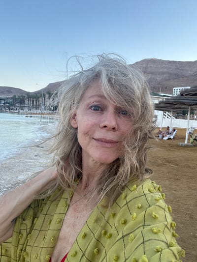 This media file is a portrait of a woman taken outdoors. She has blonde hair and blue eyes, and she is smiling. She is wearing a light green patterned shirt. In the background, there are mountains, a beach, and some buildings. The sky is clear and blue. The scene appears to be a tourist resort or a beach town. The overall mood is relaxed and happy. The image is a selfie, taken by the woman herself, as indicated by the angle of the shot and the way she is holding the camera. The location is identified as Tamar Regional Council, Israel. The time of day appears to be late afternoon or early evening, given the soft light and the long shadows. The weather is clear and sunny, with a light breeze indicated by the way her hair is blowing. There is no visible text in the image.