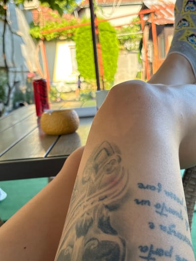 The image depicts a close-up view of a person's legs and knee, with a tattoo visible on the upper thigh. The person is wearing a sock with a Spongebob Squarepants design. In the background, a table with a red can and a small, round, yellow object is visible, along with greenery and outdoor structures suggesting a patio or garden setting. The scene appears to be outdoors on a sunny day. The specific location in Oreshak, Bulgaria, is not discernible from the image itself, but it suggests a residential or recreational outdoor space.