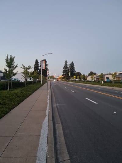 The image depicts a street scene in the city of San Carlos, United States of America, likely during dusk due to the soft, diffused light. The perspective is from a sidewalk looking out onto a multi-lane road. 

On the left side of the image, a concrete sidewalk runs parallel to a neatly manicured green hedge. Several young deciduous trees are planted along the edge of the sidewalk. Beyond the trees, parked vehicles, including a white truck and a silver van, are visible.

The road itself is paved asphalt with visible lane markings, including a double yellow line down the center and white dashed lines indicating lanes. Traffic lights are visible in the distance, some displaying red, suggesting active traffic control. A street light with a rainbow-colored banner bearing the text "CITY GOOD LIVING" is mounted on a pole near the sidewalk. Other street signs and utility poles are also present.

In the background, on the right side of the road, there are commercial buildings with modern architecture, characterized by light-colored facades and dark roofs. Some trees are planted in front of these buildings. The sky is a pale blue, transitioning towards gray, indicating the fading light of the day. No people are clearly visible in the image, and no overt actions or interactions are taking place. The scene appears to be a typical urban or suburban street environment.