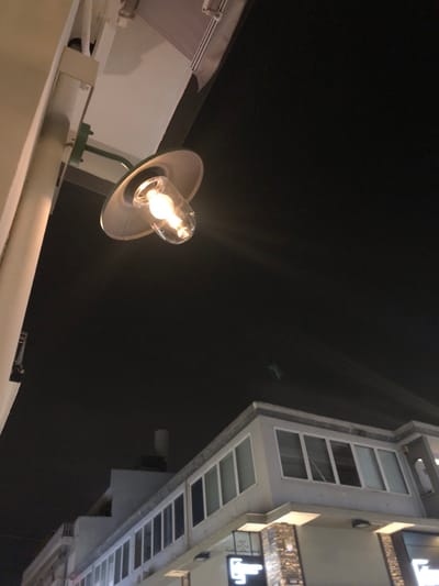 This low-angle nighttime photograph, taken in Marousi, Greece, captures a street lamp in the foreground against a dark sky and a modern building on the right.
The primary subject is a wall-mounted outdoor lamp positioned on a white ledge or structure in the lower-left to central part of the frame. The lamp features a clear incandescent bulb glowing brightly with a warm, yellowish light, creating visible starburst flares. It is housed within a clear glass enclosure and topped with a metallic, disc-shaped shade. The lamp's arm is a dark, possibly green, L-shaped support.
Above the lamp and to the right, the sky is uniformly dark, indicating nighttime. A very faint, small, dark object, possibly an aircraft or drone, is barely visible against the dark expanse in the upper-middle section of the image.
On the right side of the frame, a tall, multi-story building facade extends vertically. It features numerous rectangular windows or modular sections, arranged in a grid-like pattern. Some of these sections appear to be lit from within or by external fixtures, creating reflections and bright spots. There is an illuminated sign or decorative element on the building, but any text or specific shapes are indistinct and not clearly legible.
The overall scene is static, devoid of human activity, and the clear dark sky suggests fair weather. The artificial light sources contrast sharply with the night sky and the building's shadowy surfaces.