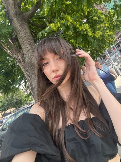 A young woman with long, dark brown hair and bangs poses for a selfie outdoors. She is wearing a black top with puffy sleeves. Her left hand, with long, dark, pointed fingernails, is visible near her hair, and she is wearing dark-rimmed sunglasses perched on top of her head. The background shows a large tree with textured bark and green leaves, and beyond that, a glimpse of a building under construction with scaffolding. In the distance, another person is visible wearing a blue shirt and jeans, with a dark bag slung over their shoulder. The scene appears to be a sunny day, suggested by the bright lighting and shadows. The overall mood is casual and relaxed. There is no visible text in the image. The location is identified as Vracar Urban Municipality, Serbia.