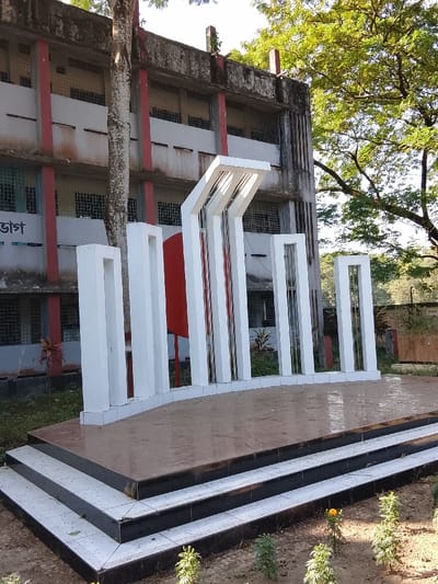 The image displays a Shaheed Minar (Martyr's Monument) situated in Shibpur, Bangladesh. The monument occupies the foreground and midground, featuring a multi-tiered base with three visible steps, constructed from white and dark-tiled surfaces. The main structure consists of several white, stylized vertical concrete pillars of varying heights, arranged in a semi-circular arc. A prominent red circular disc is centrally positioned behind the taller pillars, some of which incorporate vertical reflective metallic elements. In the background, a dilapidated two-story concrete building with red vertical supports and numerous barred windows is partially visible. A large tree with light-colored bark stands directly in front of the building. Lush green foliage from surrounding trees is present in the upper right of the frame. The immediate ground around the monument's base shows sparse vegetation, including small plants and one visible yellow flower. The scene is illuminated by natural daylight. No individuals or specific actions are present in the image.