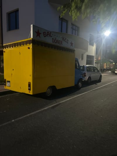 The image, rotated vertically, captures a nighttime street scene in Dzhebel, Bulgaria. A bright street light at the top left casts a strong glare, illuminating nearby foliage and the facade of a light-colored building with multiple windows.
The central subject is a vibrant yellow food truck, likely a mobile döner kebab stand, parked along the side of the road. A sign attached to its side clearly displays the text "BAL...KES DÖNER" accompanied by a red star symbol. The rear of the yellow truck shows a license plate with the registration "LK AC 7086". In front of the yellow truck, a blue pickup truck or van is parked, and further down the street, a white car is also visible.
The vehicles are situated on an asphalt road marked with a white line along its edge. The time of day is clearly night, evidenced by the darkness of the sky and the reliance on artificial lighting. The scene appears calm and devoid of people or explicit activity, suggesting it might be late evening or early morning, or simply a moment when the food truck is not actively serving. The weather appears clear.