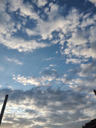 This image captures a sky filled with scattered clouds over Tervel, Bulgaria. Two dark, cylindrical poles are visible in the foreground, extending upwards from the bottom of the frame. The sky displays a mix of white and grey clouds against a backdrop of blue. In the lower right corner, a portion of a dark, tiled roof is visible, suggesting the presence of a building. A small, dark silhouette of a person can be seen near the edge of the roof. The lighting suggests it is either early morning or late afternoon, with the sun creating highlights on the clouds.