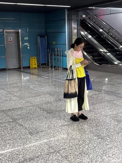 The image shows a woman standing indoors, presumably in a public space. She is wearing a light-colored coat, a yellow top, black pants, and black shoes. She is holding a cell phone and appears to be looking at it. A purse hangs from her shoulder.

In the background, there's an escalator and a blue-tiled wall with some signs on it. A yellow caution sign is also visible. The floor is gray and appears to be made of concrete or stone.

Based on the location 龙溪街道, China, it's likely this scene is from a subway station or a similar public transportation hub.