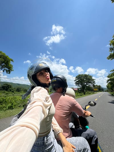 In Hàm Thuận Bắc, Vietnam, on a sunny day with scattered clouds, two individuals are riding a motorcycle on a paved road. The person in the front, a woman wearing a helmet with a clear visor and a light-colored sheer blouse over a tank top and jeans, is capturing a selfie. She is puckering her lips towards the camera. Behind her, a person wearing a pink t-shirt and a black helmet is driving the motorcycle. A third person, wearing a light-colored helmet, is visible behind the driver. The road stretches ahead, flanked by lush green vegetation and trees, with hills in the background. The scene appears to be a scenic route, likely in a rural or semi-rural area within Hàm Thuận Bắc.