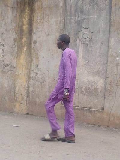 A man in a purple traditional outfit walks along a paved area beside a textured concrete wall. He wears sandals and has wrist adornments. The wall behind him is characterized by discoloration and a decorative circular element. The light is overcast, creating soft shadows and muted colors.