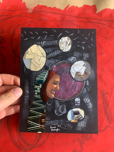 The image shows a piece of abstract art, possibly a collage or mixed media piece, set against a vibrant red background with some brown paper elements at the top. The artwork itself is on black paper and features a profile view of a smiling person wearing a head covering made of patterned fabric. Several circular collages of paper scraps are placed around the central figure, some appearing to depict abstract shapes or fragmented imagery. There is also a large, roughly circular, reddish-purple shape in the center of the black paper. White chalk-like marks and lines are drawn across the black background, adding to the abstract and somewhat chaotic feel of the piece. In the bottom right corner of the artwork, there is a handwritten signature and date: "Pavel 2017 Kuragin". The artwork is held by a person's hand, which is visible on the left side of the frame. The overall impression is of a creative and expressive artwork. There is no identifiable location in the city of Sergeevo, Russia, depicted in this image.