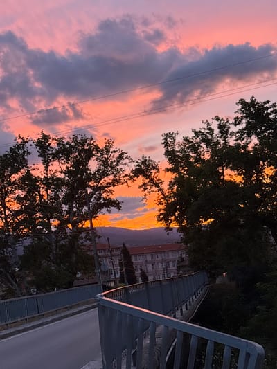 A vertical, medium-shot, full-frame photograph captures a sunset over the city of Mikrevo, Bulgaria. The sky is filled with a gradient of warm colors, from a soft pink at the top to vibrant orange and yellow hues closer to the horizon. Dark, wispy clouds are scattered across the sky, adding texture and depth. In the foreground, silhouetted trees with dense foliage frame the scene on either side. A modern pedestrian bridge with a gray metal railing extends from the bottom left corner towards the center of the frame, crossing over a roadway. The roadway itself is paved and slopes gently upwards. In the middle ground, a building with a red tiled roof and multiple windows is visible, hinting at a residential or institutional structure. Beyond this, a range of distant hills is faintly discernible against the colorful sky. The overall impression is one of a peaceful evening in a Bulgarian town.