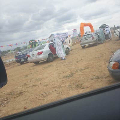 In Wuro Ngangandi, Nigeria, an outdoor event is taking place under a cloudy sky. The scene is characterized by a dirt ground, several parked cars, and a large orange inflatable archway. A banner reading "GROUND BREAKING AND COMMISSIONING" is visible, suggesting the event's purpose. Several individuals, some dressed in traditional white and light blue attire, are interacting near a silver car with its door open. One person is holding a golden staff. Other cars, including a blue Peugeot and a silver Toyota, are parked in the vicinity. Colorful flags are strung across a fence in the background, adding to the festive atmosphere.
