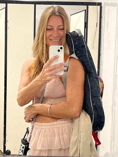 A woman with blonde hair and fair skin takes a mirror selfie in a room that appears to be a dressing room or bathroom. She is wearing a light pink lace bralette and a matching pink ruffled skirt. She is holding a white smartphone in her right hand, angled to capture her reflection. A black bag with a zebra print strap is visible hanging from her left shoulder, and she is also carrying a dark-colored bag slung over her right shoulder, partially obscuring her arm. The background consists of tiled walls and a framed mirror, suggesting an indoor setting. The lighting is bright and even. The room exhibits features consistent with a changing room or perhaps a hotel bathroom in Katunayake, Sri Lanka.