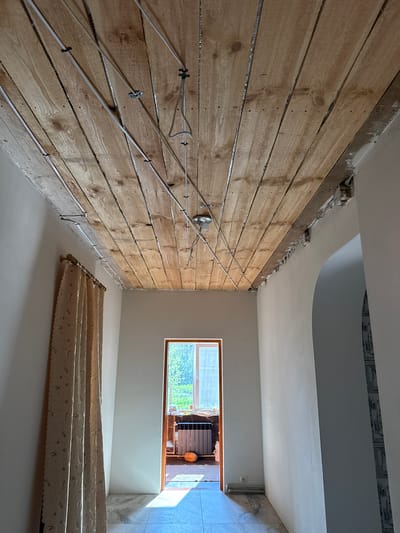 This media file captures an interior view of a room undergoing renovation, likely in Luka, Ukraine. The primary subject is a wall clad in horizontal wooden planks, with visible electrical wiring neatly secured to it. The wiring runs diagonally across the wall, connected to clips and leading to a single light fixture. The wall's rough, unfinished nature suggests it's in a preparatory stage of construction or decoration.

Above the wooden wall, a portion of the ceiling is visible, featuring a curved architectural element and some exposed structural details, hinting at ongoing work. The room also includes a window with a wooden frame, offering a glimpse of greenery outside, suggesting a daytime scene with natural light filtering in. Below the window, a patterned curtain is draped, adding a contrasting element of domesticity to the otherwise utilitarian setting. The floor appears to be a light-colored, smooth surface, possibly concrete or a similar material.

The overall scene depicts a room in transition, with evidence of electrical work in progress. The lighting suggests it is daytime, and the view through the window implies an outdoor environment consistent with a residential area. There is no visible text in the image. The atmosphere is one of active renovation rather than a lived-in space.