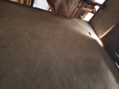 An interior view of a partially constructed or dilapidated building in Gindin Dutse, Nigeria, shows a rough, earthen-colored wall that occupies the majority of the frame. The wall has an uneven texture with visible pores and small imperfections. In the upper portion of the image, exposed wooden beams of the roof structure are visible, along with what appears to be a tarpaulin draped over some of the framing, suggesting unfinished work or a protective covering. Natural light enters from openings in the roof, casting shadows and highlighting the textured surface of the wall. To the right, a dark wooden frame suggests the presence of a doorway or window, with a sliver of bright light emanating from its edge. A small, dark, rectangular object, possibly a nail or a loose piece of debris, is embedded in the wall. The overall impression is one of an unfinished or neglected structure.