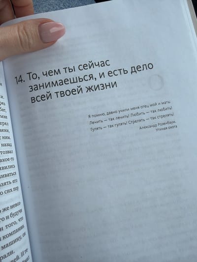 A female's left hand with manicured nails and light pink gel polish holds an open book, displaying Russian text on white pages. The prominent text on the right page is numbered "14" and reads, "То, чем ты сейчас занимаешься, и есть дело всей твоей жизни" (What you are doing now is the business of your whole life). Below this, a smaller quote is visible, attributed to Alexander Rozenbaum from "Утиная охота" (Duck Hunting): "Я помню, давно учили меня отец мой и мать: Лечить — так лечить! Любить — так любить! Гулять — так гулять! Стрелять — так стрелять!" (I remember, my father and mother taught me long ago: To treat - so treat! To love - so love! To walk - so walk! To shoot - so shoot!). Additional smaller Russian text appears on the opposing left page and subtly below the main quote on the right. A fragment of blue denim fabric is visible in the upper left background, indicating the presence of a person wearing denim, likely seated. The lighting is soft and even, suggesting an indoor or sheltered environment. The scene takes place in Yus'ki, Russia, though the specific details of the location within the city are not visually discernible.