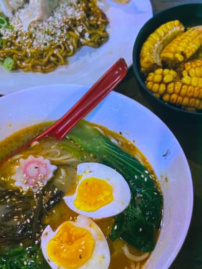The image shows a close-up view of several dishes of food, possibly at a restaurant. In the foreground, a white bowl contains a ramen soup with noodles, broth, a narutomaki (fish cake), green leafy vegetables, and two halves of a hard-boiled egg. A red spoon is resting in the bowl. To the right, a dark bowl holds several pieces of corn on the cob. In the background, a white plate displays a serving of noodles with green onions and what appears to be some sort of topping. The lighting is somewhat dim, and the image has a cool, slightly desaturated tone. The specific location in Cadek, Indonesia cannot be determined from the image.