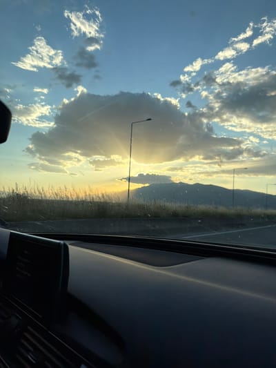 From the interior of a car, the shot captures a partly cloudy sky in Sidirokastro, Greece. The sun is visible behind the clouds and illuminates mountains on the horizon. Streetlights line the highway. There are raindrops on the car window. The dark car interior frames the shot.