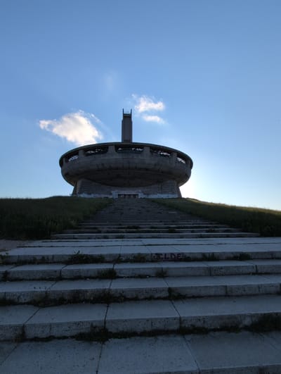 The image shows the Buzludzha Monument, a former communist party house in Shipka, Bulgaria. The monument, an imposing Brutalist structure with a distinctive flying saucer-like shape, is situated on a hilltop. A grand staircase, constructed from concrete and showing signs of wear and tear with grass growing between the steps, leads up to the monument. The monument itself is weathered and appears abandoned, with graffiti visible on its lower section. A tall, slender tower rises from the center of the main circular structure. The sky above is a clear blue, with a single white cloud and several small dark specks, possibly birds, in the distance. The surrounding landscape is a grassy slope, partially illuminated by the sunlight. The overall impression is one of a monumental structure in a state of decay, set against a serene natural backdrop.