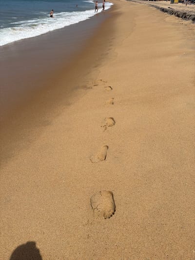 The image depicts a sandy beach with the ocean in the background. Footprints are visible in the sand, leading away from the water. There are several people on the beach, some wading in the water and others standing on the sand. The setting appears to be a tropical beach, likely in Arugam Bay, Sri Lanka, known for its surfing spots and relaxed atmosphere. The sun is shining, casting shadows on the sand. The overall scene suggests a pleasant day at the beach.