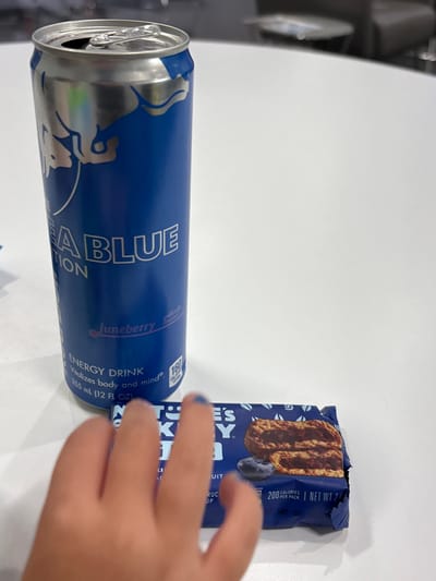 The image displays a blue Red Bull Juneberry Edition energy drink can and a Nature Valley Blueberry flavored granola bar on a white surface. The can is open, with its silver top visible. The granola bar is in its packaging. The fingers of a hand with blue nail polish are visible near the granola bar. The drink can states "ENERGY DRINK Vaolizes body and mind", contains 355ml, and has 160 calories. The granola bar indicates it has 200 calories per pack.