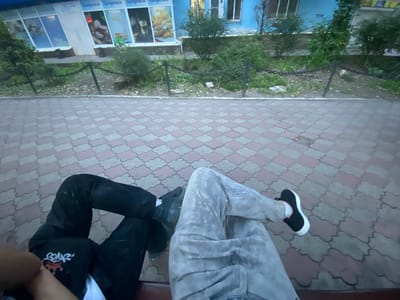 The image shows a first-person perspective of two individuals sitting on a bench in a city environment. The individuals are only visible from the waist down, showing their legs and feet. One individual wears black pants and a black shirt with a white graphic. The other individual wears light-colored, possibly gray or beige, pants and black sneakers with white soles.

The setting appears to be outdoors, with a paved walkway made of interlocking tiles in shades of pink and gray. Behind the walkway, there is a low, black metal chain fence. Beyond the fence, there is an area with greenery, including bushes and trees. In the background, a building with a blue facade and windows is visible. The windows have images displayed in them, some of which appear to be food items. The overall environment suggests an urban or semi-urban public space, possibly a park or sidewalk in front of a business. The lighting suggests it might be late afternoon or early evening, as there are shadows cast on the pavement. The image does not contain any overt signs or clues that definitively place it in Antratsyt, Ukraine, but the context of the query implies this is the intended location.