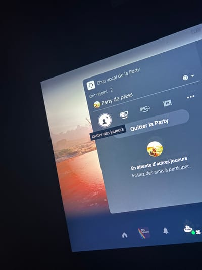 This image captures a digital screen displaying a gaming console interface, likely a PlayStation, within an indoor environment in Illkirch-Graffenstaden, France. The screen is angled, showing a semi-transparent dark blue overlay menu on the right. This menu, titled "Chat vocal de la Party" (Party voice chat), indicates "Ont rejoint : 2" (Joined: 2) and specifies the group as "Party de press" (Press Party). Visible interactive elements include buttons labeled "Inviter des joueurs" (Invite players) and "Quitter la Party" (Leave Party). A message states, "En attente d'autres joueurs Invite des amis à participer." (Waiting for other players Invite friends to participate.). A circular profile icon depicts a figure on an animal within a landscape scene. The background behind the menu shows a digital landscape featuring reddish-orange water, rock formations, and what appears to be a desert environment. The bottom edge of the screen displays a row of interface icons: a home symbol, an application icon with "ABC builders" and a rainbow logo, a notification bell, and a user icon showing a green status dot and the number "26". The surrounding area outside the screen is dark, indicating a dim room.