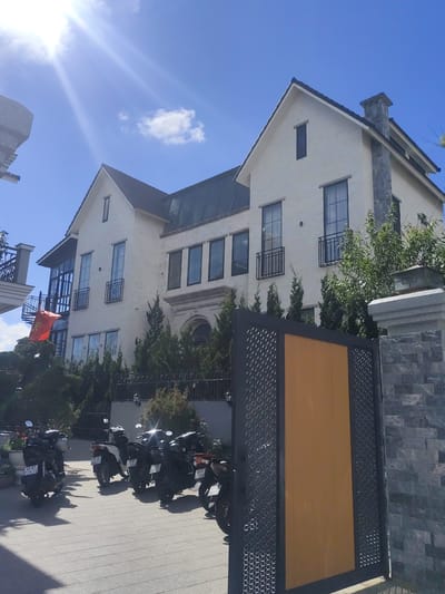 The image shows a modern, two-story building with a light-colored facade, multiple windows with black frames, and a dark tiled roof. The building features a prominent stone-clad chimney. In front of the building, a bright orange gate with a decorative metal lattice is visible, flanked by a stone wall. A small courtyard or driveway in front of the gate is paved with light-colored tiles, and several motorcycles are parked there. A red flag with a yellow star, presumably the Vietnamese flag, is visible hanging from a balcony on the left side of the building. Green trees and bushes are planted around the property, and the sky is clear with a bright sun casting rays. The scene is located in the Lam Vien area of Da Lat, Vietnam, as indicated by the architectural style and the presence of the Vietnamese flag.