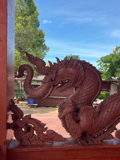 The image shows a close-up of a detailed, carved wooden sculpture of a mythical serpent-like creature, possibly a Naga, rendered in a deep reddish-brown hue. The creature's head is prominent, with an elongated snout, open mouth revealing teeth, and intricate swirling patterns adorning its crest and body. The sculpture is positioned in the foreground, with an out-of-focus background showing a courtyard area with red brick paving, a few buildings with tiled roofs, green trees, and a bright blue sky with scattered clouds. A blue plastic bin sits on a small bench in the middle ground. This appears to be an exterior architectural element, possibly at a temple or cultural site in Baan Hua Thanon, Thailand, given the style of the carving and the tropical vegetation.