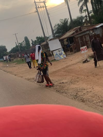 This image, set in Agban, Nigeria, captures an outdoor street scene, likely during late afternoon or early morning, characterized by soft, diffused light and a low sun visible in the upper left. The setting features a paved road on the right and a wider, dusty unpaved path on the left, lined with rudimentary structures made of corrugated iron and wood, possibly indicating small shops or informal stalls.
The main subject is a person, likely a street vendor, standing by a small display cart on the edge of the paved road. They are wearing a colorful, patterned short-sleeved top (featuring green, red, and yellow hues), dark shorts, and distinct red open-toed sandals. A white cloth is draped over their head and shoulders. The cart beside them appears to hold various goods, though they are not clearly discernible.
Several other people are visible further down the dirt path, suggesting ongoing street activity. These include individuals in a red shirt and yellow pants, and another in a light blue/grey shirt. In the background, palm trees and other foliage are visible, and numerous utility poles with crisscrossing power lines span the sky.
A notable detail is a white sign with red lettering that reads "PUSS," mounted on a pole near one of the corrugated iron structures. The immediate foreground on the right side is partially obscured by a large, blurry red object. The scene portrays a typical, active roadside environment.
