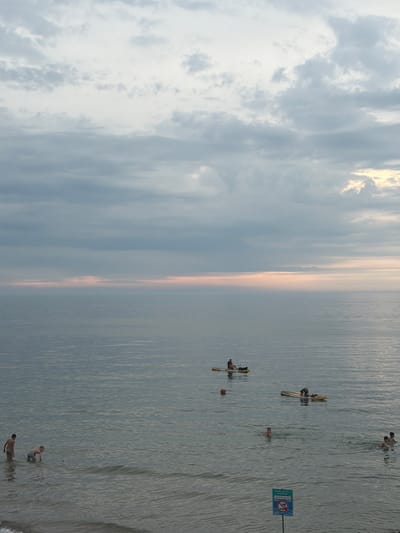 The image depicts a body of water, likely the Baltic Sea, with several people engaged in various water activities. In the foreground, two individuals are partially submerged in the water. Further out, a person is on a paddleboard, and another is in a kayak. The sky is overcast with clouds, and a hint of pink can be seen along the horizon, suggesting it might be dawn or dusk. The overall scene conveys a sense of a calm and recreational atmosphere. Given the context of Zelenogradsk, Russia, this scene is likely situated on the Zelenogradsk beach or a nearby coastal area.