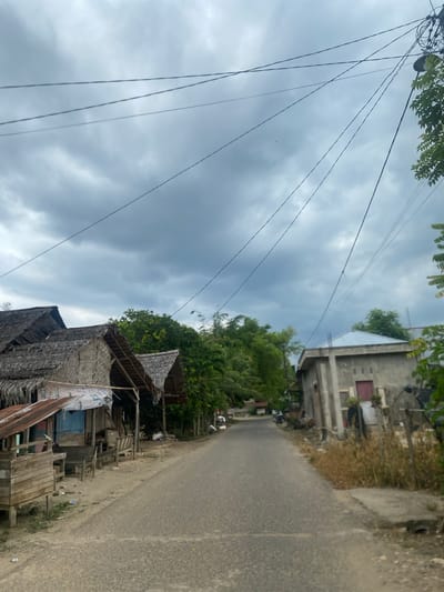 The image depicts a street in Kebonbaru, Indonesia. The street is paved and appears narrow. On one side of the street are buildings of varying construction styles. The closest building has a thatched roof and wooden supports. Further down the street is a more modern concrete building with a red door. Lush green vegetation is visible. Overhead, electrical wires stretch across the sky, which is filled with dark, cloudy skies.