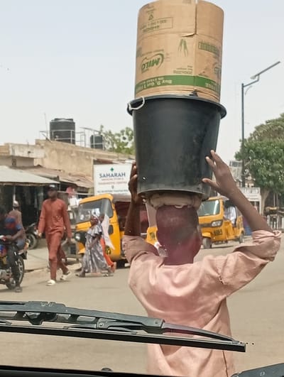 The image depicts a daytime street scene in Hotoro North, Kano, Nigeria, viewed from inside a vehicle with windshield wipers visible at the bottom. The central subject, seen from behind, is a person wearing a light-colored long-sleeved garment, balancing a black plastic bucket topped with a large brown cardboard box on their head. A light-colored cloth cushion is positioned between the head and the bucket. The cardboard box displays "MILO" branding and text including "200X20G" and "Buy 9, Get 1 Free." The person's arms are raised, hands lightly touching the load for stabilization.
In the midground, a paved road shows pedestrian and vehicular activity. A male in a reddish-brown tunic and trousers with a dark cap and white shoes walks left-to-right. Two other pedestrians, one in a patterned dress and another partially obscured, walk away from the viewer. Multiple yellow three-wheeled auto-rickshaws are present, one marked with "6965" on its rear. A motorcycle is partially visible on the far left.
The background features low-rise buildings with flat roofs, several topped with black water tanks. A white sign with green lettering clearly identifies "SAHARAH MEDICINE STORE & DIAGNOSTIC CENTRE NO. 138 HOTORO NORTH / TANIMBAR ROAD KANO." Sparse trees with green foliage and a modern street light pole are also visible. The sky is bright and hazy.