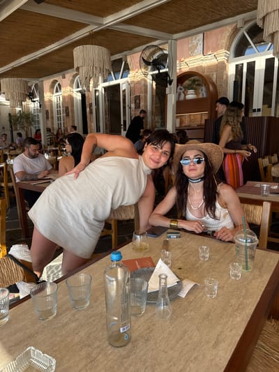 Two women are seated at a table in an outdoor restaurant in Chania, Greece. The woman on the left is leaning forward with her left hand on the table and her right arm behind her back. She is wearing a white sleeveless dress. The woman on the right is wearing a straw hat, sunglasses, and a white top. She has a necklace and bracelets on. In front of them on the table are several glasses, a bottle of water, a small box, and a menu. The restaurant has a thatched roof and stone walls. Other patrons are visible in the background.